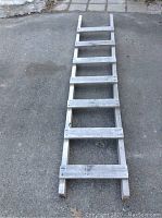 Single wooden straight ladder, 6 wide rungs, grey weathered finish shown laying on ground outdoors.