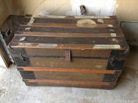 Front view of an antique wooden slat trunk with metal reinforcements, showing wear, scratches, and a latch closure.