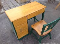 Full view of maple desk with natural finish top, green frame, and matching chair with maple seat and green backrest and legs shown on pavement.