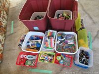 Photo showing two large red plastic totes with lids, several white bins and small baskets filled with various pens, pencils, paper clips, and other small office and school supplies.