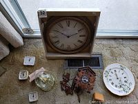 Photo of all clocks together including wall clock in box, wooden cuckoo clock with carved leaves, Royal Albert china plate clock, quartz 400 day clock, and four small alarm clocks.