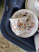 Close-up of a china cup with portraits of Prince Charles and Princess Diana and matching decorated plate beneath, with cherub and gold trimming details.