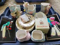 Overall shot of Royal Doulton Bunnykins toddler dishware and vintage baby room decor showing plates, bowl, cup, silver spoons, boxed Bunnykins item, ceramic animal holders, figurine, and framed textile art.