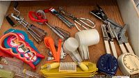 Various kitchen utensils including manual egg beaters, measuring spoons, and cups arranged in a drawer, showing the range of items and materials.