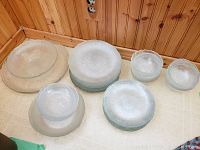 Stacks of glass dishware including large plates, small plates, bowls, and platters shown on a table against a wood paneled wall.