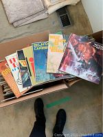 Two cardboard boxes filled with assorted vintage 1950s vinyl records. Visible record covers include "Pennies from Heaven," "Belafonte Caribbean," and "What About Us." Records are stacked and partially arranged, showing colorful mid-century graphic design.