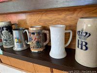 Eleven beer steins displayed on wood shelf in two rows, variety of styles and materials visible