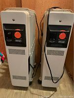 Front view of two white DeLonghi portable oil-filled radiator heaters side-by-side with control panels visible and power cords looped.