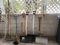 Full view of various gardening and snow removal tools lined up against a wall showing multiple handles and the tops of the tools.