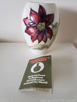 Front view of the oval Moorcroft Pottery vase with red flower and pamphlet in front.