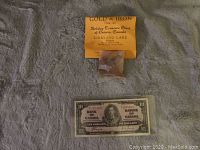 Photo showing the gold and iron packet with the orange informational card from Holiday Treasure Chest above a 1937 Bank of Canada ten dollar bill.