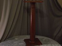 Front view of the wooden plant stand showing its cylindrical column and square base and top, under soft lighting and neutral background.
