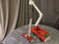 Two vintage metal fire truck toys displayed on a round surface with gray cloth backdrop, larger Tonka with white bucket crane and smaller Super Tiger with ladder.