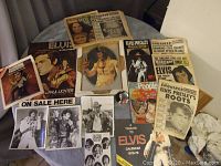 Photos showing collection of Elvis Presley memorabilia spread out on a round table, including newspapers, posters, calendars, and magazines with visible wear.