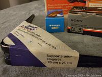 Close-up photo showing L-shaped black shelf support brackets with paper label and small AC adapters boxed (Sanyo and Sony brand).