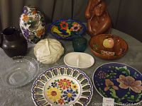 Photo shows assortment of serving dishes including ceramic plates with floral motifs, a clear glass divided dish, a dark pottery pitcher, a large decorated urn, wooden bowl with fruit motif, a white ceramic bowl, a green glass dish, and a carved wooden sculpture.