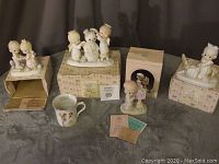 Photo showing five Precious Moments figurines arranged on and next to boxes, along with a coffee mug and printed materials.