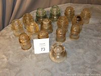 Seventeen glass insulators grouped on a grey cloth surface showing variety in shape and color, mostly amber, some clear and greenish.