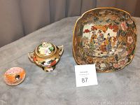 Three Asian-designed dishes including a large bowl, small sugar dish with lid, and small pin dish displayed on grey fabric.