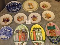 Lot of 11 assorted serving plates and dishes featuring fruit motifs, blue and white scenic designs, and colorful village scenes displayed on a grey surface.