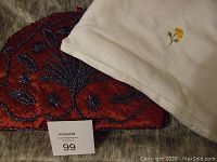 Two tea pot cozies placed side by side on a surface. One is white with embroidered yellow flower, the other red silk with black bead details.