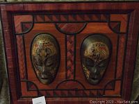 Photo showing both handcrafted wooden masks mounted on a woven wooden frame with geometric detailing and some visible wear.