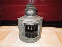 Front view of the galvanized tin marine signal lamp with red fresnel glass lens on port side, showing lamp top chimney vent and aged metal body.