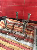 Pair of silverplated candleholders with twisted connecting stems, arranged on a striped cloth in front of a red tile background.