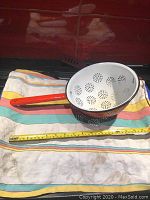Vintage enameled metal strainer placed on a cloth with measuring tape along its length, showing red exterior, white interior, and perforated circular patterns for straining.