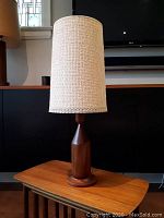 Full view of the mid-century modern teak lamp with woven fabric shade on a wooden surface, showing overall shape and size.