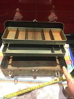 Opened vintage green metal tackle box with two interior metal lifting trays, top tray segmented into compartments, showing signs of rust and wear.