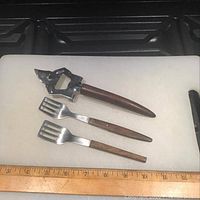 Photo showing two wooden-handled herring lifters and a wooden-handled bottle opener arranged on a white cutting board with a ruler showing measurements.