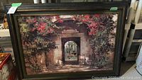 Full view of the framed painting showing a doorway surrounded by red and green foliage.