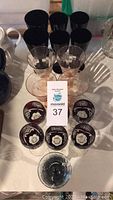 Black wine glasses in two sizes, red cut glass liqueur glasses with floral patterns, and pink stemmed wine glasses arranged on a table.