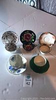 Photo showing six assorted tea cups and matching saucers arranged in pairs on a white surface. Various floral designs and gold trimming detail visible.