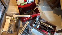 Wide view of red metal toolbox open with various hand tools and hardware inside, including hammer, saw, and Jobmate wrench set.