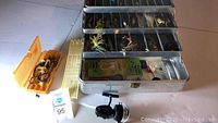 Metal fishing tackle box open with multiple trays filled with various fishing lures and a fishing reel beside it.
