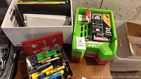 General view of office supplies including pens, highlighters, scissors in a red Hudson's Bay cookie tin, cardboard box with notebooks, and plastic trays with additional office items.