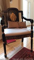 Front view of the wooden chair showing the upholstered seat, carved back with gold leaf motif, and padded bolster pillow.