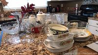 Lot contents including glass punchbowl set, coffee cups, CorningWare dishes, flatware, and ceramic plates on kitchen counter