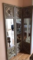 Three-panel wooden folding screen with vertical mirrors and decorative carved floral panels at top and bottom, standing folded against wall.