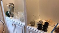 Overview of white cabinet top holding glassware including glass vases, serving platter and ceramic coffee mugs in black and white along with conch shell on side cabinet