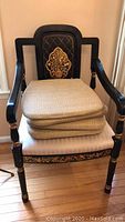 Front view showing black wooden chair with carved and painted gold floral design on backrest. Three beige seat cushions stacked on upholstered white seat.