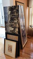 Grouped view of the framed art pieces stacked against a wall, including a tall cityscape photo and smaller framed paintings