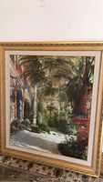 Framed art showing garden or patio scene with plants, trees, columns and brickwork on carpeted floor near white wall.