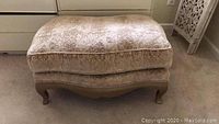 Front side view of rectangular upholstered ottoman showing beige patterned fabric and wooden carved cabriole legs.