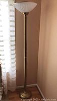 Full view of the 72 inch tall gold colored floor lamp positioned next to a window with curtains.