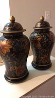 Two black ceramic urns with lids showing leaf and flower gold patterns