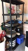 Black plastic shelving unit filled with various kitchen and household items such as bakeware and pitchers.