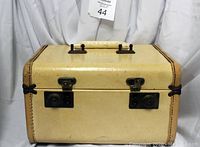 Closed beige rectangular travel case with two locking clasps on the front, beige handle on top, stitched edges, and black corner protectors.